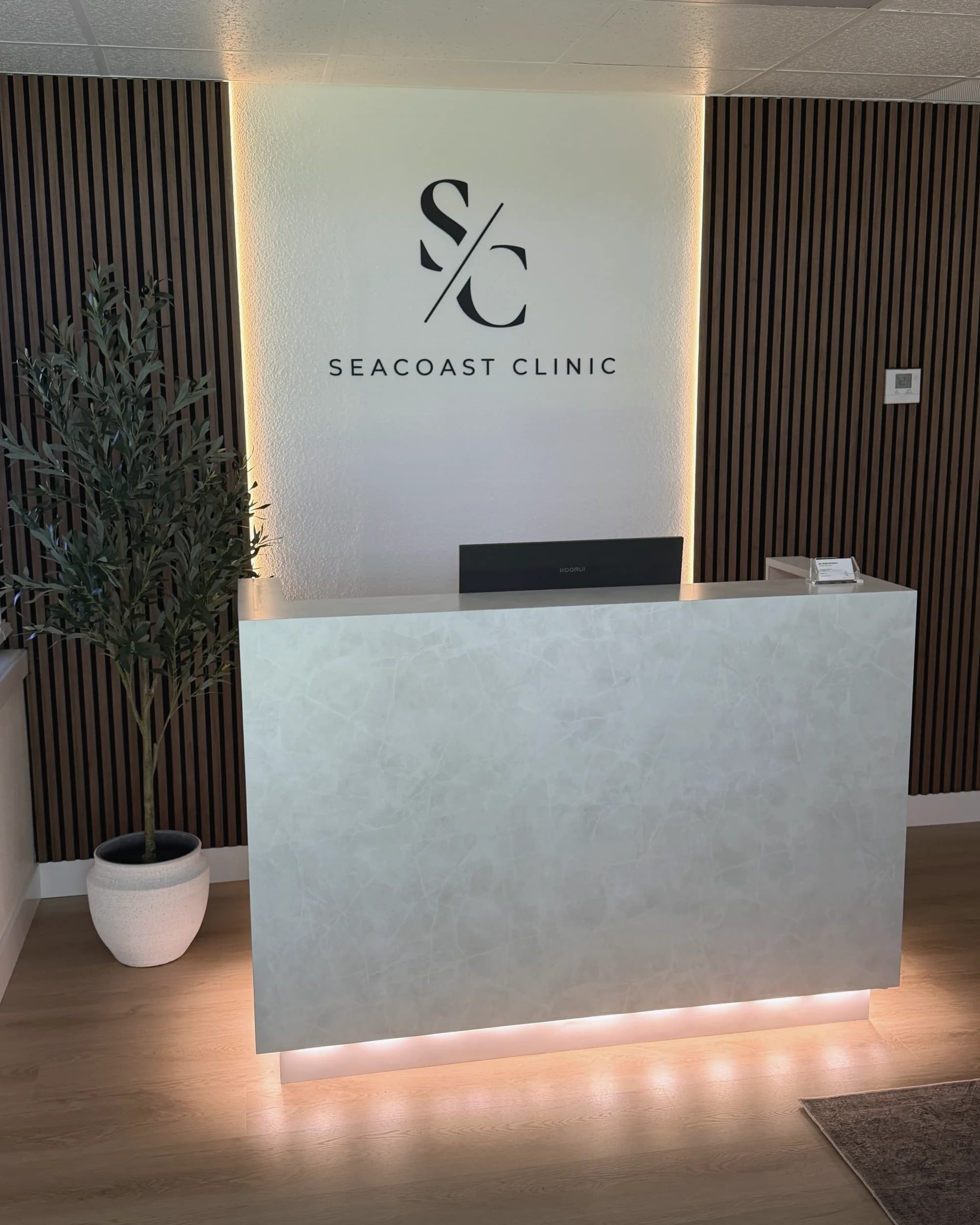 The front desk at SeaCoast Clinic beneath the backlit Sea Coast sign, its pale stone counter warmed by golden light